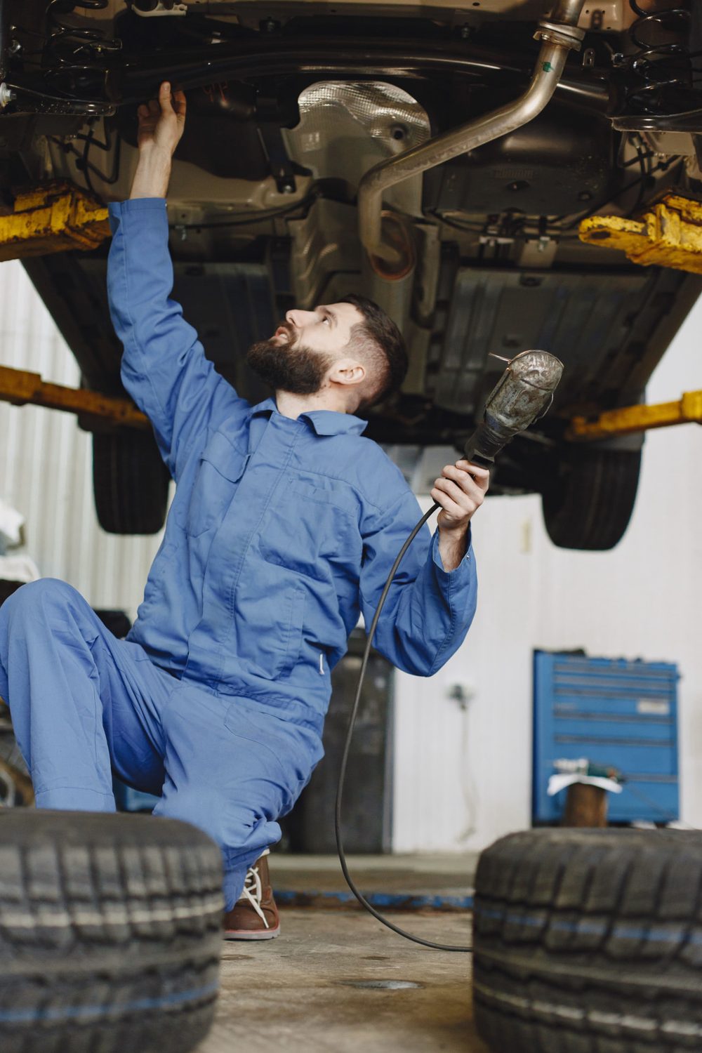 Technician in blue coveralls kneeling beneath a vehicle raised on a shop lift, holding a work light up to the undercarriage
